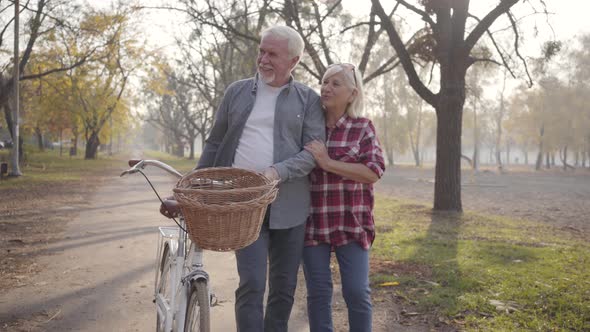 Happy Senior Caucasian Couple Walking with Bicycle Along the Alley in the Foggy Park. Retired Mature