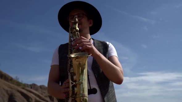 Musician Is Dressed in Hat and Waistcoat and Playing Saxophone at Nature at Summer Day