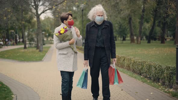 Wide Shot of Senior Couple in Covid19 Face Masks Walking with Bouquet of Flowers and Shopping Bags