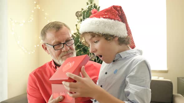 Grandpa Wearing a Santa Hat Makes a Christmas Present To His Teenage Grandson. The Boy Is Delighted