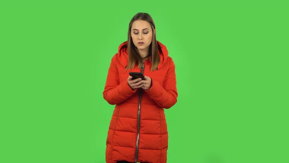 Lovely Girl in a Red Down Jacket Is Texting on Her Phone. Green Screen