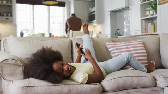 African american daughter lying on couch laughing at tv with father in background