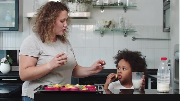 White Mother with Her Black Mixed Daughter Eating Cupcakes with Milk in the Kitchen