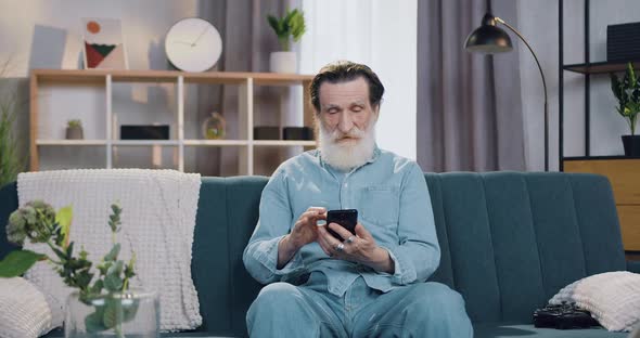 Bearded Man which Sitting on Soft Couch at Cozy House and Uses Smartphone to Make Call