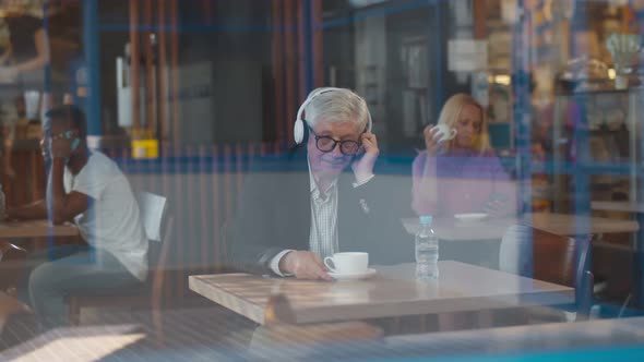 View Through Window of Mature Man with Headphones at Table in Cafe