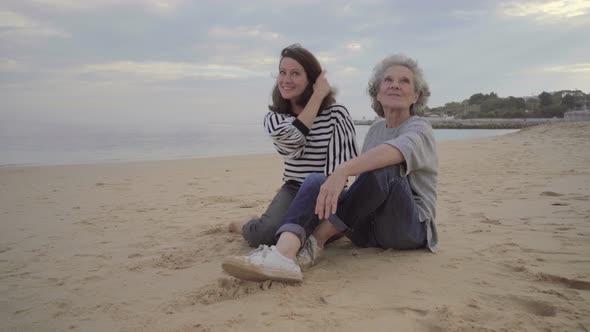 Happy Old Mother Hugging and Kissing Beautiful Adult Daughter on Sand Near Sea