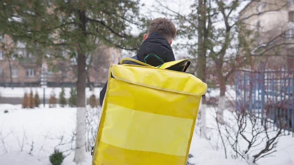 Back View of Young Man Hanging Food Delivery Backpack and Walking Away on Winter Day Outdoors