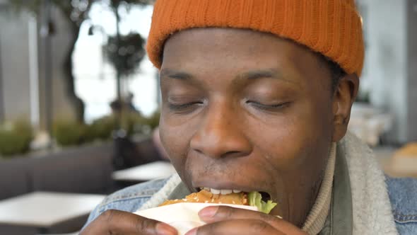 Black Guy in Jacket and Hat Tastes Delicious Burger in Cafe