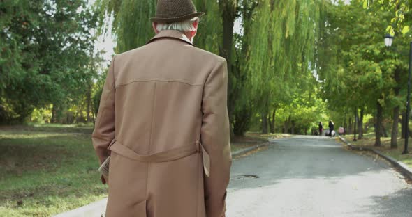 Back Look of Alone Grandfather in Outerwear Walking with Crutch Slowly on Square