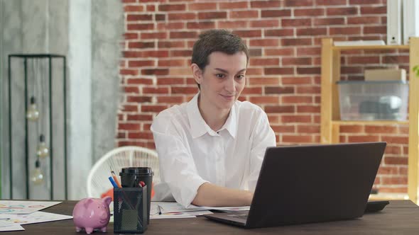 Cute businesswoman prints on laptop smiling and sitting at an office table. Short-haired woman works