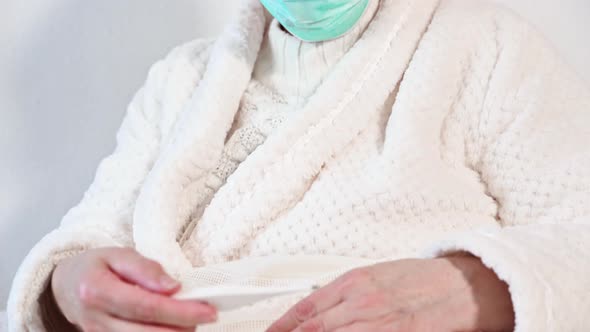 Coronavirus. Sick Senior Woman with Protective Mask Holding Thermometer Sit on Bed, Upset Old Mature