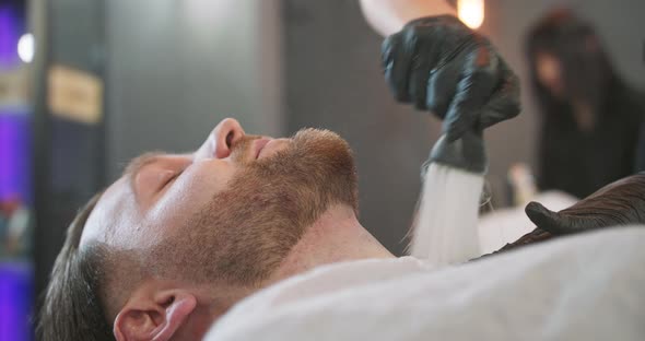 Closeup Head of Young Bearded Man is Sitting on the Barber's Chair with Cape and Hands of Female