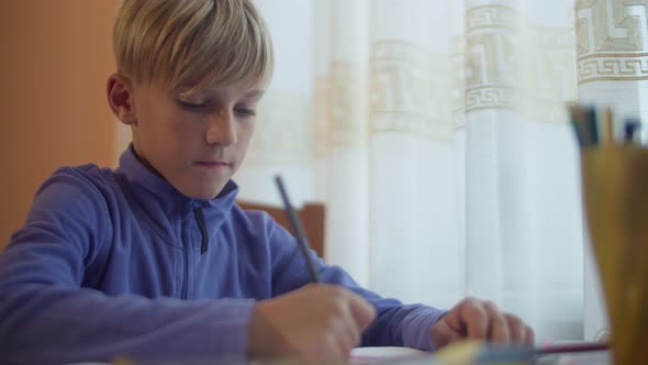 A schoolboy at the window draws at the table with pencils. Portrait of a drawing child