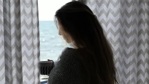 Young Woman in a Blanket and with a Cup of Coffee on the Background of Curtains and the Sea