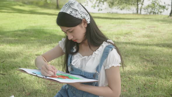 Young asian woman sitting drawing picture with colorful paint brush while leisure in the park.