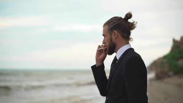 Young Guy in Business Suit with Tied Hair Thoughtfully Watching in Distance at Sea and Smoking
