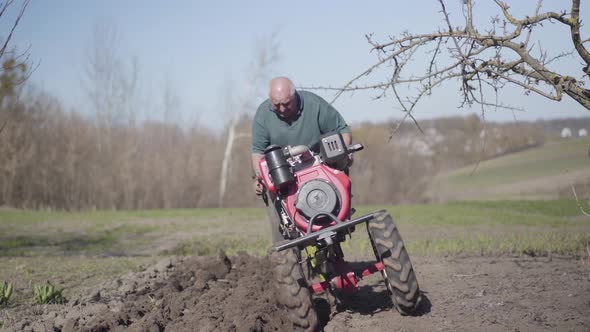 Portrait of Serious Elderly Caucasian Man Walking Along Rural Garden with Furrower. Old Farmer