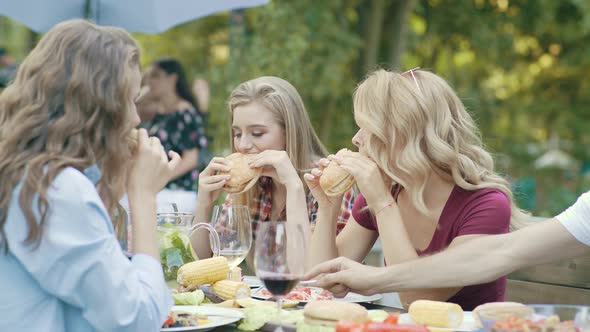 Friends Eating Burgers Sitting At Dinner Table At Outdoor Party