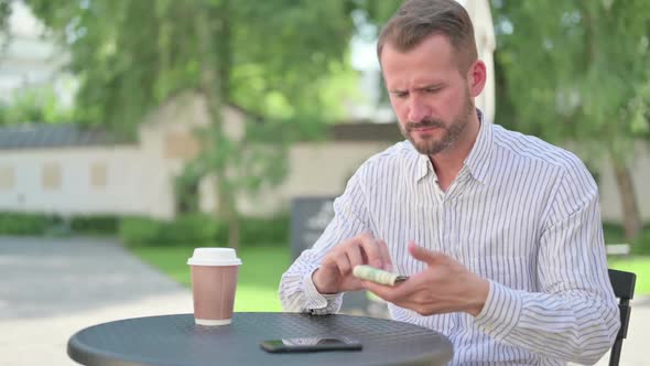 Middle Aged Man Feeling Sad While Counting Dollars in Outdoor Cafe