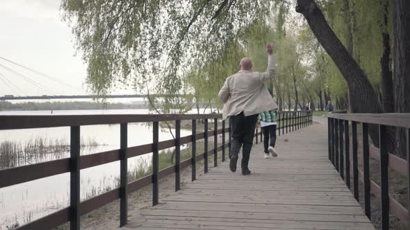 Little Boy Running Away From His Grandfather on the Wooden Bridge Near the River