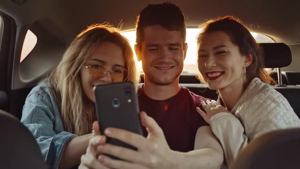 Handsome Young Guy Making Selfie with Pretty Girls on Phone or Smartphone in Back Seat of Moving Car