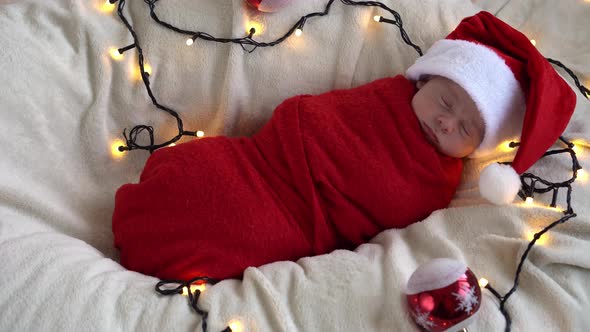 Top View Portrait First Days Of Life Newborn Cute Funny Sleeping Baby In Santa Hat Wrapped In Red