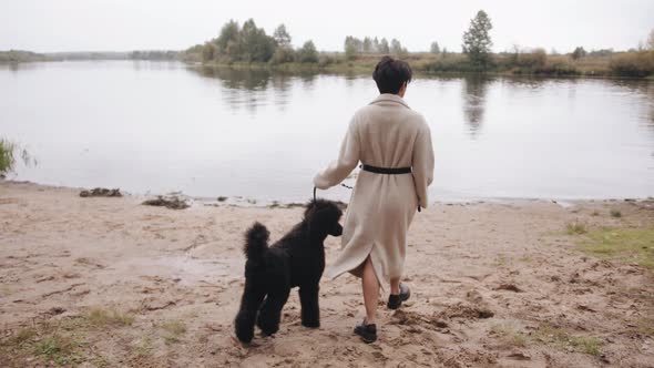 A Young Brunette in a White Coat Goes to the River and Holds a Black Poodle on a Leash