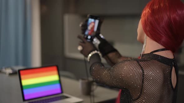 Proud Travesty Woman Taking Selfie on Smartphone with Laptop Standing on Table with LGBT Flag Screen