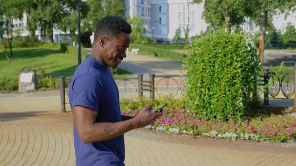 Smiling Afroamerican Man Walks Street Uses Mobile Phone Holding Cup with Coffee