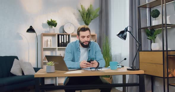 Guy Sitting at His Workplace at Home and Uses His Mobile to Read News or Write Message