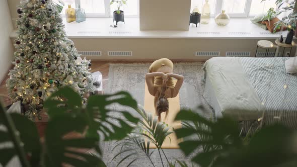 A Woman Does Yoga at Home in a Room with a Bed and a Christmas Tree in the Background