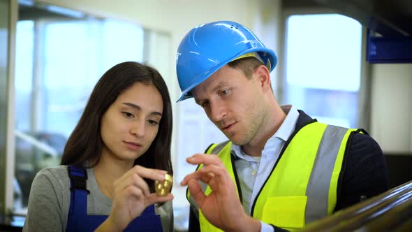 Female skilled worker and coworker controlling metal part in factory