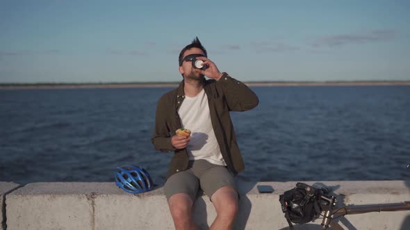 Male Cyclist Eats Sandwich and Drinks Coffee to Go While Sitting on Promenade By Sea