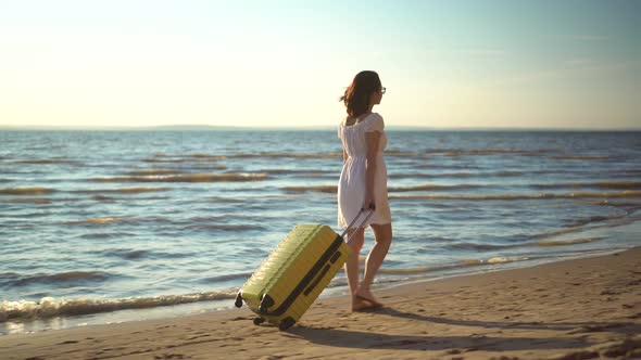 Young Woman Walks Along the Beach of the Sea with a Yellow Suitcase. A Girl in a White Dress Walks
