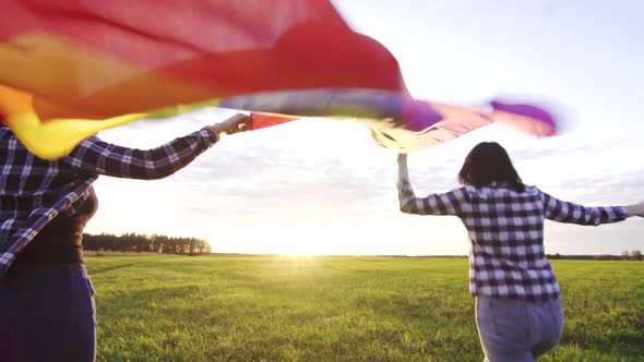 Two Young Lesbian Girls in Shirts and Glasses Run Across the Field at Sunset with an LGBT Flag