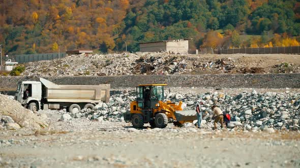 a group of three men loads stones near the river into bucket of an excavator.