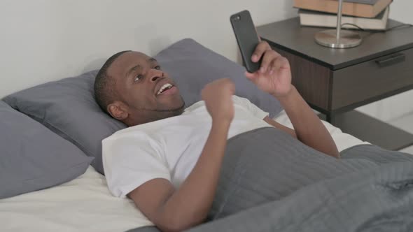 African Man Celebrating Success on Smartphone in Bed