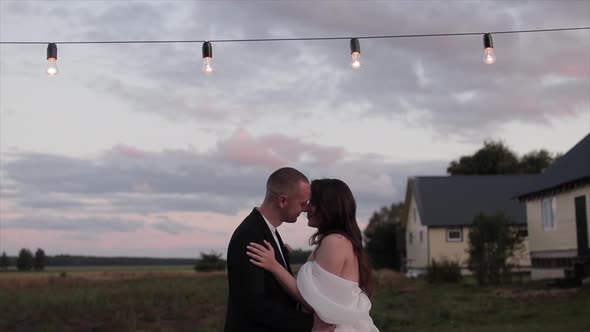 The Bride and Groom Stand Facing Each Other and Hugging on the Lawn Near the Houses in the Village