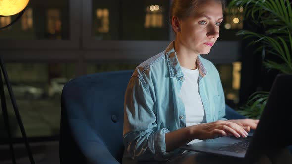 Woman is Sitting in the Armchair and Working on a Laptop at Night or Texting Someone