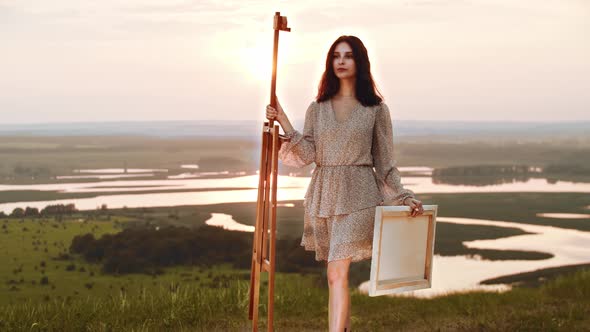 A Young Woman Artist Walks to an Easel Standing on the Sunset Field and Putting a Canvas on It