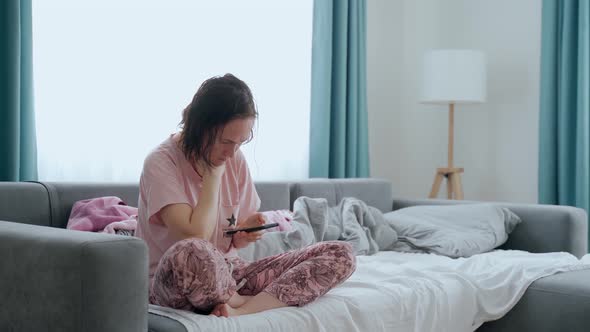 A Woman in Pajamas Sits on a Sofa with a Phone
