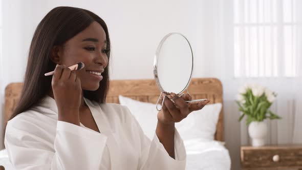 Black Woman Making Makeup Using Cosmetic Brush Sitting In Bedroom