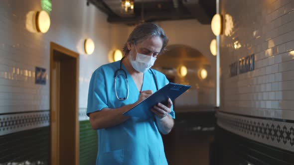 Senior Surgeon in Medical Mask Writing on Clipboard in Corridor at Hospital