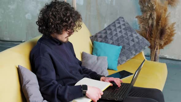 Young Man is Browsing at His Laptop and Writing Notice on His Paper Notebook