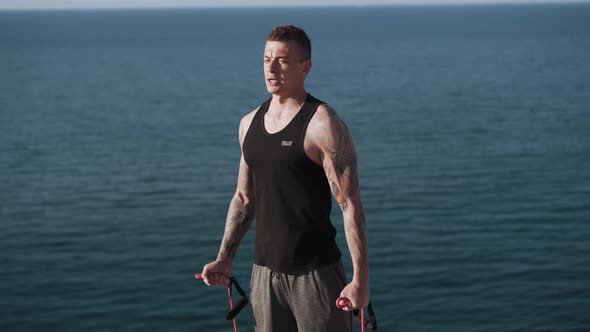 Muscular Tattooed Man Working Out with Elastic Bands Outdoor with Sea Views