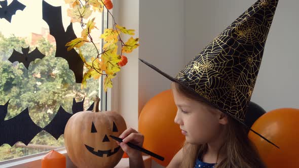 Preparation for Halloween Little Girl Paint Funny Face on Pumpkin in Witch Hat with Black Marker