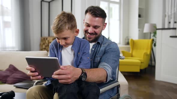 Man in Wheelchair Playing Video Game on Tablet PC Together with His Cheerful 8-Aged Son