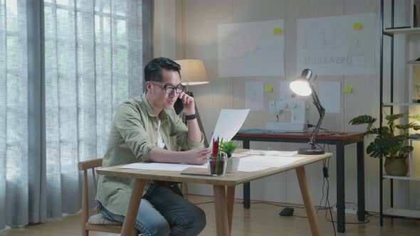 Asian Man Designer Talking On Smartphone While Looking At The Layout Bond In Hand At The Office