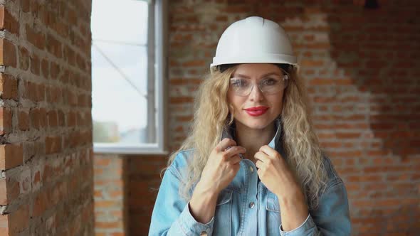 A Young Woman in a Work Helmet and Denim Clothes and Goggles Stands at the Construction Site