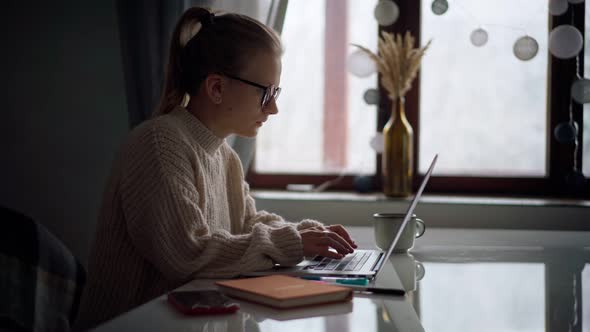 A Young Girl Wearing Glasses Typing on a Laptop and Making Notes in a Notebook